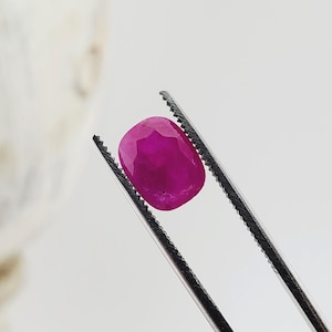 May include: A vibrant, cushion-cut ruby gemstone with a deep magenta-red hue and multifaceted surface, held securely by a pair of silver-toned metal tweezers with serrated tips, showcasing the stone's clarity and polished, reflective gemstone facets.