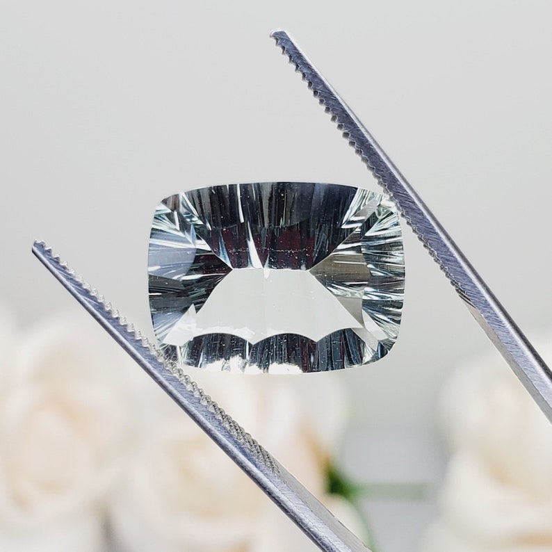 May include: A clear, faceted, cushion-shaped gemstone with a light green hue. The gemstone is held by tweezers against a white background.