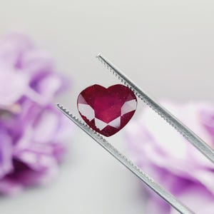 May include: A heart-shaped, faceted ruby gemstone held by silver tweezers. The ruby is a deep red color with multiple facets that catch the light. The background is blurred, with hints of purple flowers.