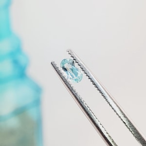 Puede incluir: Una pequeña piedra preciosa de color azul claro y corte ovalado sostenida por unas pinzas de metal plateado. La piedra presenta una superficie facetada que refleja la luz, mostrando un color cian pálido translúcido y un aspecto pulido.