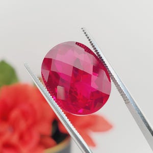 May include: A vibrant red oval-shaped gemstone with a faceted cut, held by silver tweezers.