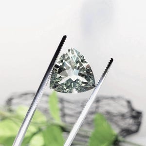 May include: A triangular green gemstone with facets, held by tweezers.