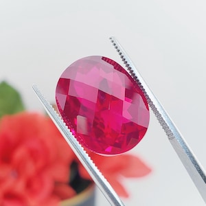 May include: A vibrant, oval-shaped, faceted gemstone in a deep magenta-red color. The gemstone is held securely by a pair of silver-toned metal tweezers, showcasing its reflective, multi-faceted surface and brilliant, saturated red hue.