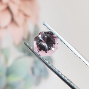 May include: A faceted, light pink gemstone held by a pair of silver tweezers. The gem has a geometric shape with multiple facets that reflect light. The background is blurred, with hints of pink and green.