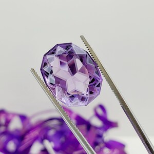 May include: A large, faceted, purple amethyst gemstone held by tweezers. The gemstone is oval-shaped and has a high degree of clarity.