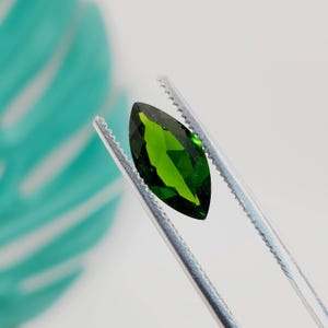 May include: A marquise-cut, vibrant green gemstone is held by silver tweezers against a white background. The gem's facets reflect light, showcasing its clarity and rich color. The tweezers are in sharp focus, highlighting the gem's shape and brilliance.