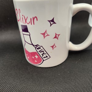 May include: White ceramic mug with a pink and purple design. The mug features a bottle with a pink liquid and a tag that says "Elixir". There are also pink and purple stars on the mug.
