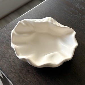 May include: A white ceramic bowl with an irregular, organic shape that mimics the appearance of a natural shell. The bowl features a smooth, glossy finish with deep, undulating folds and a wide, open center, creating a sculptural look.