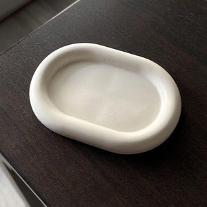 May include: An oval-shaped tray with a smooth, matte, off-white finish. The tray features a raised, rounded rim and a shallow, recessed center, presenting a clean, solid construction with a uniform, neutral color throughout the entire piece.