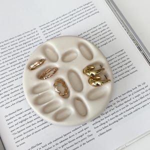 May include: A round, off-white ceramic jewelry dish with multiple oval indentations, holding three thin gold-toned rings and one pair of smooth, curved gold-toned hoop earrings, resting on an open book with printed text.