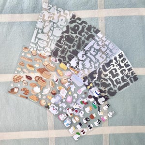Puede incluir: Una colección de hojas de pegatinas transparentes con varios diseños. Las pegatinas incluyen siluetas de gatos, alimentos como pan y pasteles, y lindos personajes de dibujos animados. Las pegatinas son en tonos de blanco, gris y negro.