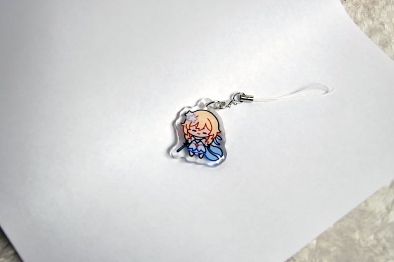 May include: A clear acrylic charm depicting a cartoon character with blonde hair, blue and white clothing, and a small wand. The charm has a silver-coloured clasp and a white lanyard. The design is a chibi-style illustration.