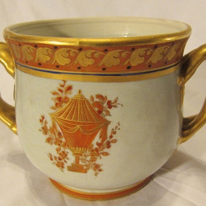 May include: A white porcelain cup with two gold handles. The cup has a gold rim and a gold and orange floral design. The cup is decorated with a gold and orange urn.