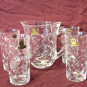 May include: A set of five clear glass pieces, including a pitcher and four glasses. The glasses and pitcher have a cut glass design and feature a gold label with the text "RCR Oneida".