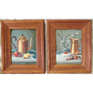 May include: Two framed still life paintings. The first painting features a copper teapot, apples, and lemons on a table with a blue tablecloth. The second painting features a copper pitcher with a sprig of white flowers on a table with a blue tablecloth.