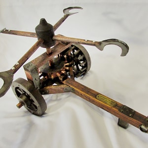 May include: A wooden model of Leonardo da Vinci's war machine, the 'Carro da Vinci', with a metal frame and four metal hooks. The model is on a white surface and has a brass plaque that reads 'LEONARDO DA VINCI - CARRO BALLA - 1500 - PARIS'.