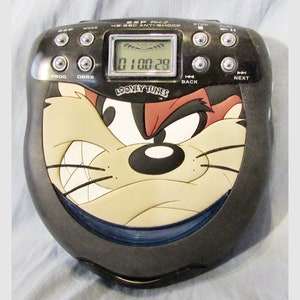 May include: A black and blue portable CD player with a cartoon image of Tasmanian Devil from Looney Tunes. The player has a digital display showing "01 00:28" and buttons for play, stop, next, back, mode, prog, and dbbs.