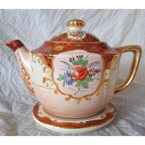 May include: A vintage porcelain teapot with a matching saucer. The teapot features a floral design with red, orange, and blue flowers on a cream background. Gold trim accents the edges and handle. The lid is also decorated with floral patterns.
