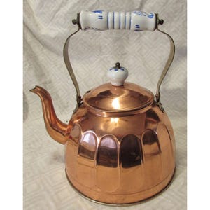 May include: A copper tea kettle with a white and blue ceramic handle and lid knob. The kettle has a polished finish and a curved spout. The handle is attached to the kettle with metal brackets.