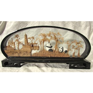 May include: A decorative diorama featuring a detailed landscape with a pagoda, trees, and two pandas. The scene is encased in a black, oval-shaped frame with a black base. The pandas are black and white.