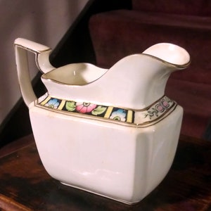 May include: White ceramic creamer with a gold trim and floral design. The creamer has a square base and a rounded spout.