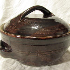 May include: A dark brown glazed ceramic casserole dish with a matching lid. The round pot features two side handles and a central handle on the lid. The surface has a glossy finish with subtle horizontal grooves and etched cross-hatch markings.