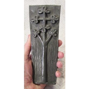 May include: A dark gray rectangular wall plaque with a tree design. The tree has a trunk and branches with four-leaf clover-shaped leaves. The plaque is held in a hand.