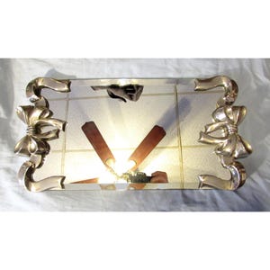 May include: A rectangular silver-toned mirror with decorative bow accents on each corner. The mirror has a grid pattern and two brown, angled supports. The overall design is vintage and elegant.