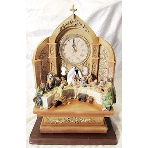 May include: A decorative quartz clock featuring a sculpted scene of the Last Supper at the base. The clock face has Roman numerals and is set within an arched, tan-colored frame topped with a cross. The figures are painted in muted tones.