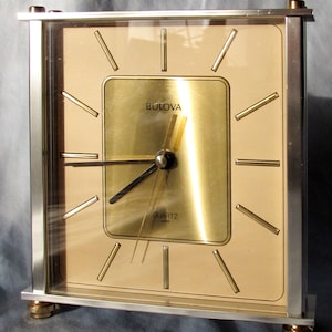 May include: A silver and gold-toned Bulova quartz clock with a square face and black hands. The clock has a light brown background with gold-colored hour markers.