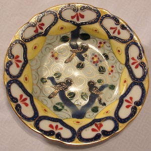 May include: A small, white porcelain saucer with a scalloped edge. The saucer is decorated with a blue and gold floral pattern and two birds in flight. The birds are painted in shades of blue, green, and yellow.
