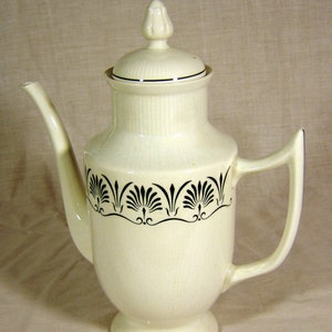 May include: Cream-colored ceramic coffee pot with a lid and a handle. The pot features a black floral design around the middle and a decorative finial on the lid. The pot has a spout for pouring.