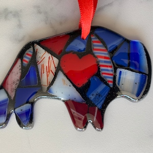 May include: A colorful, mosaic-style ornament in the shape of a bison. The ornament features red, white, and blue glass pieces, with a red heart in the center. A red ribbon is attached for hanging. The bison is approximately 4 inches long.