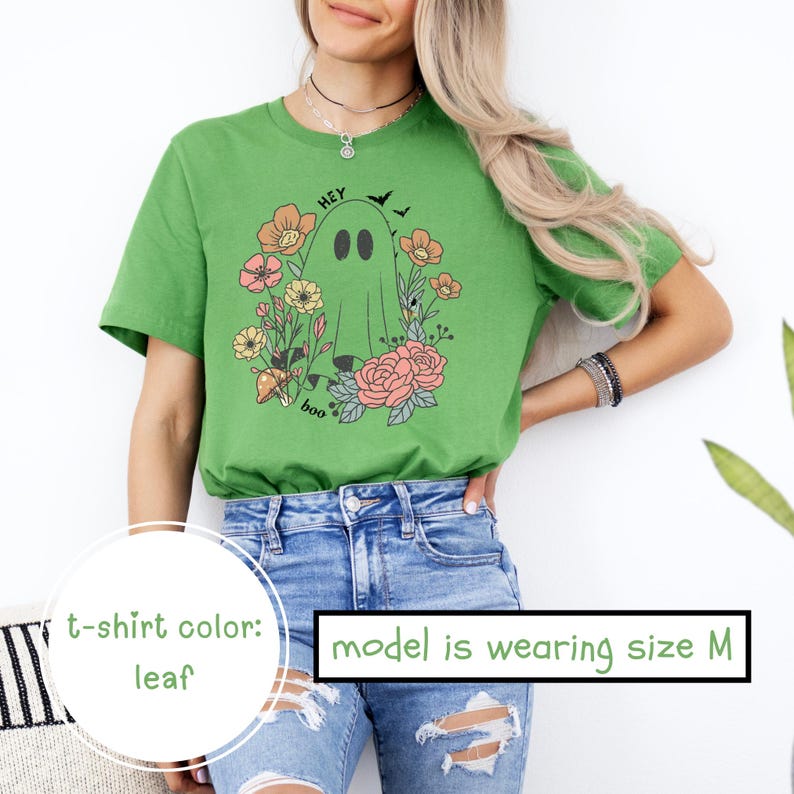 May include: A green t-shirt with a floral design and a ghost graphic. The text "Hey Boo" is printed on the shirt. The model is wearing size M.