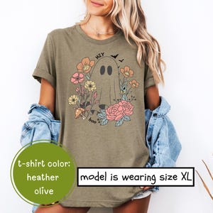 May include: A heather olive t-shirt with a graphic of a ghost with the text "Hey Boo" and floral accents. The model is wearing size XL.