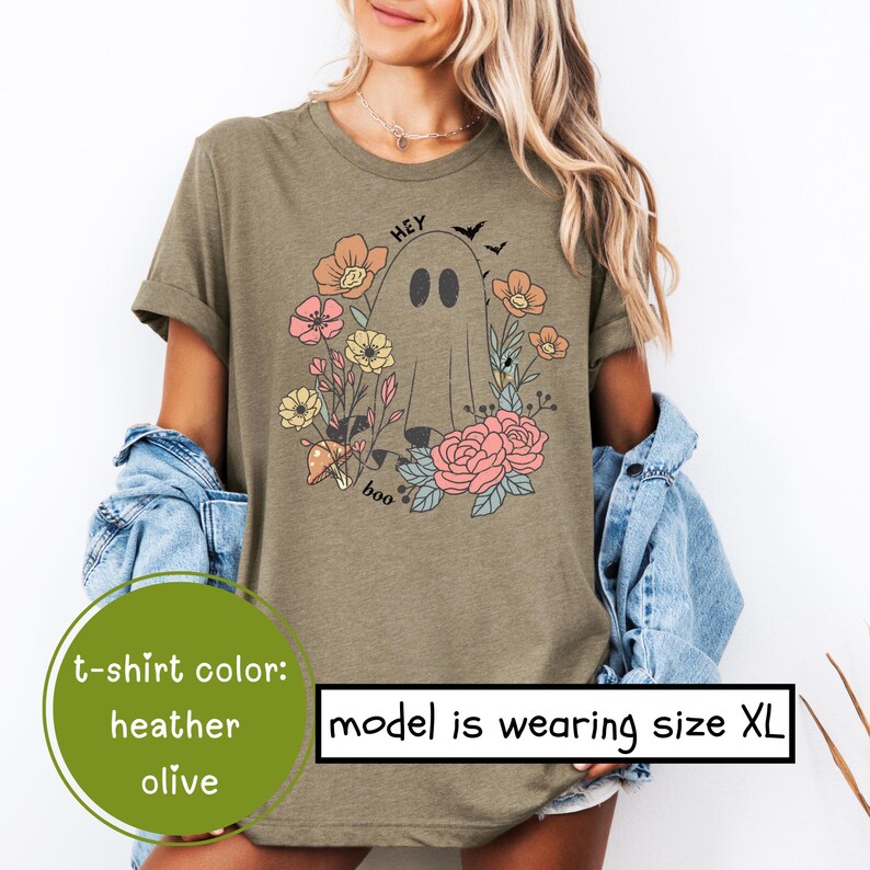 May include: A heather olive t-shirt with a graphic of a ghost with the text "Hey Boo" and floral accents. The model is wearing size XL.