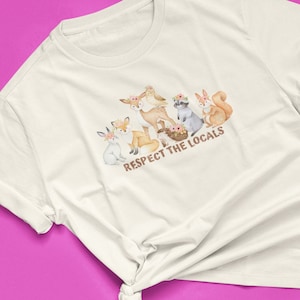 May include: White t-shirt featuring a watercolour illustration of woodland animals wearing flower crowns. The text "Respect the Locals" is printed below the animals.