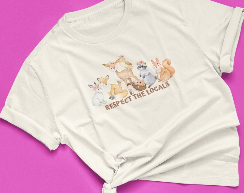 May include: White t-shirt featuring a watercolour illustration of woodland animals wearing flower crowns. The text "Respect the Locals" is printed below the animals.