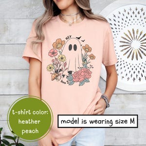 May include: A heather peach t-shirt with a graphic of a ghost with the text "Hey Boo" and floral accents. The model is wearing size M.