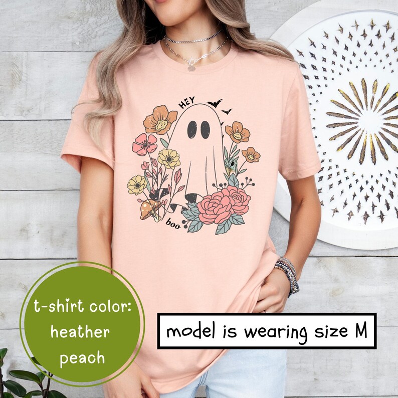 May include: A heather peach t-shirt with a graphic of a ghost with the text "Hey Boo" and floral accents. The model is wearing size M.