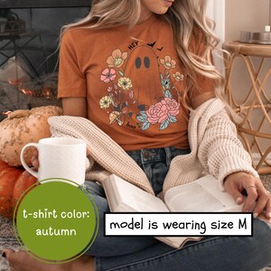 May include: A woman wearing a brown t-shirt with a ghost and floral design. The shirt says "Hey boo".