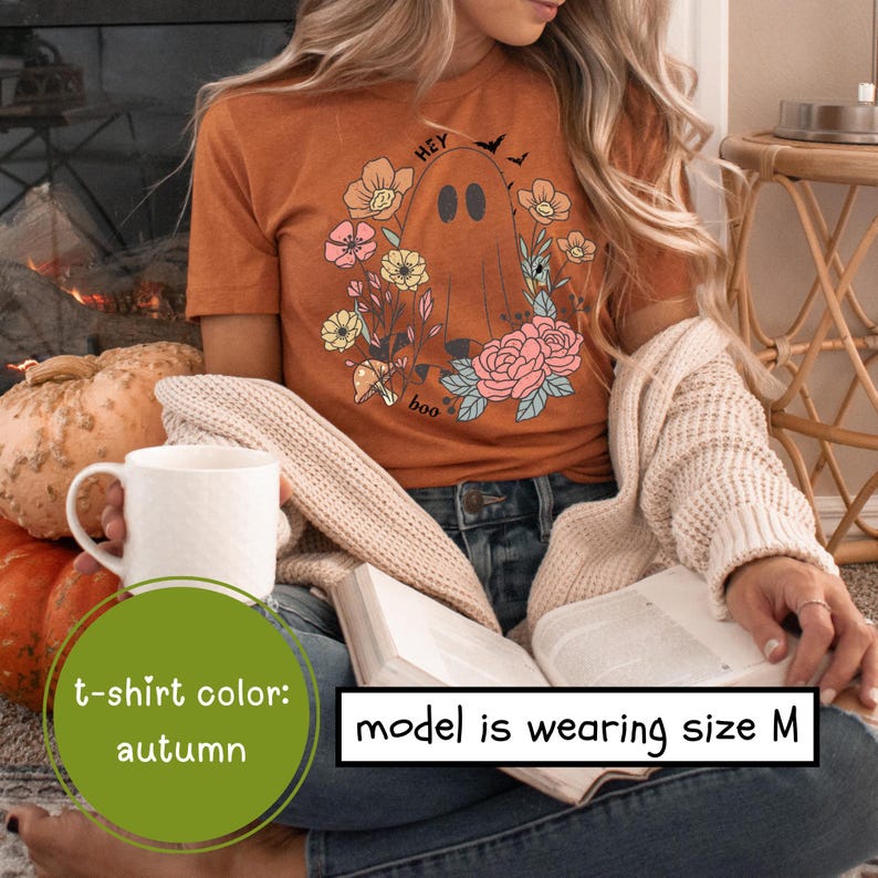 May include: A woman wearing a brown t-shirt with a ghost and floral design. The shirt says "Hey boo".