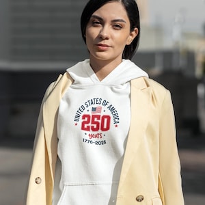 May include: A white hooded sweatshirt with the text "UNITED STATES OF AMERICA" encircling a design with the number "250" and the American flag. The sweatshirt also includes the years "1776-2026". The person is wearing a light yellow blazer.