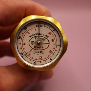 May include: A vintage gold-coloured gauge with a white face and red markings. The gauge reads "Waters Mfg. Inc. Tokoy Watch Gauge". The gauge is held in a hand against a pink background.