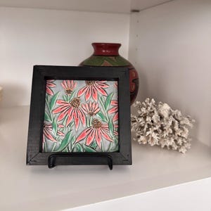 May include: A framed decorative tile featuring a floral design with pink and green flowers on a gray background. The tile is displayed on a black metal stand, set against a white shelf with a decorative vase and coral.