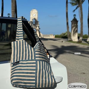 May include: A blue and beige striped tote bag with a patchwork design. The bag has a long strap and wooden bead accents. The brand name "Colemico" is visible on a small black tag. The bag is leaning against a white car.