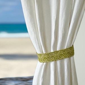 May include: White linen curtains are held back by a green patterned tieback. The tieback is a band of woven fabric with a geometric design. The background shows a beach and ocean scene.