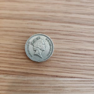 Op de afbeelding: Een zilveren munt met een profiel van een gekroond hoofd, waarschijnlijk een monarch, met tekst eromheen. De munt ligt op een houten achtergrond. De details van de munt zijn scherp.