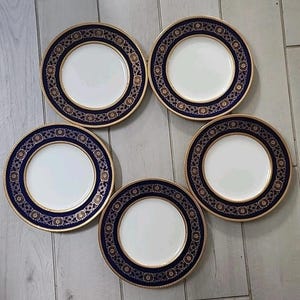 May include: A set of five round dinner plates featuring a white centre and a wide dark blue border. The border is decorated with a repeating gold floral pattern and finished with a thin gold rim along the outer and inner edges of the plate.