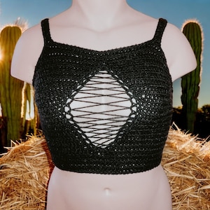 May include: Black crochet crop top with a diamond-shaped cutout and crisscross detailing. The top has thin straps and a textured appearance. The top is displayed on a mannequin against a desert backdrop.